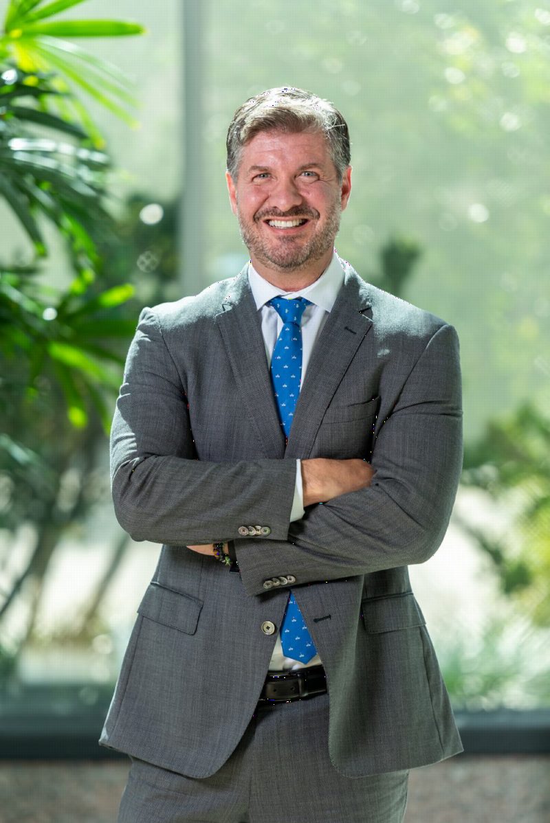 Gui Gatti, de terno cinza e gravata azul, sorrindo de braços cruzados